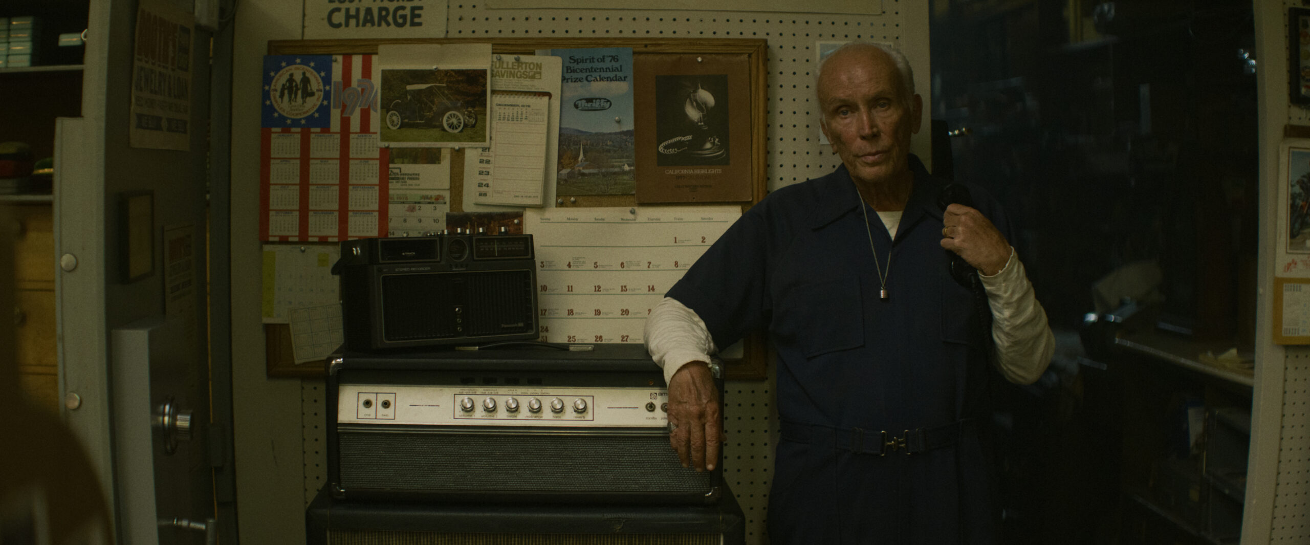 "Older man in blue coveralls stands in a dimly lit workshop, resting on an amplifier, with a radio, calendar, and various tools and papers on the pegboard wall behind him, suggesting a repair or electronics shop environment."
