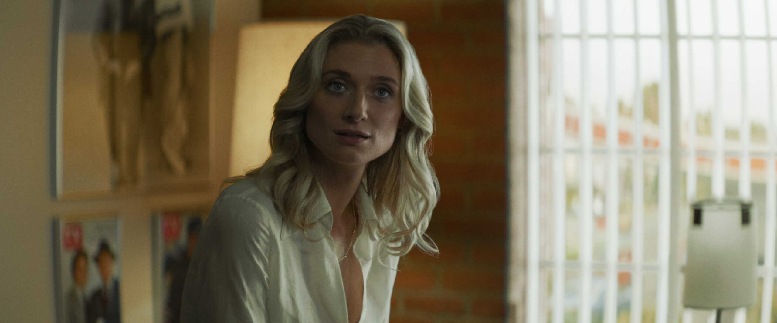 "A woman with blonde hair sits indoors near a lamp and large window with vertical bars. The room has a brick wall, framed pictures, and soft lighting, giving a cozy, modern atmosphere."