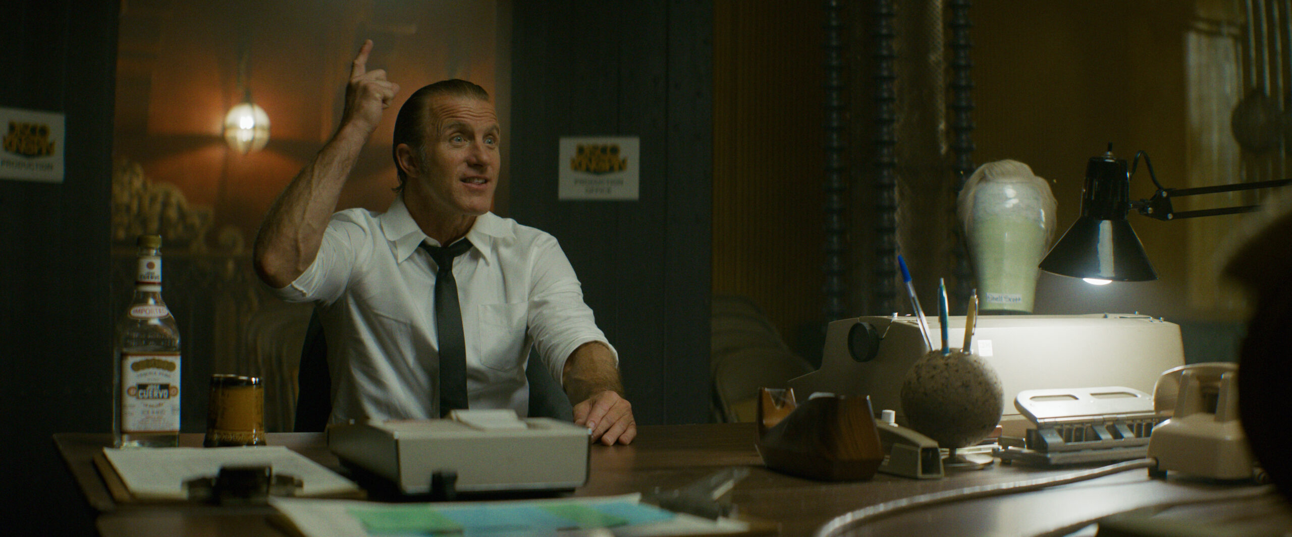 "Man in a white shirt and tie sits at a cluttered office desk with papers, a drink, and a bottle, gesturing energetically. The office is dimly lit with vintage decor, a typewriter, desk lamp, and industrial pipes in the background."