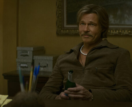 "A man with a mustache sits at a desk in a warmly lit office, holding a green bottle. The room has a lamp, filing boxes, and papers, with a painting and organized supplies in the background."