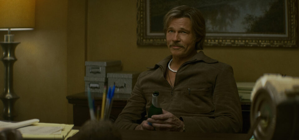 "A man with a mustache sits at a desk in a warmly lit office, holding a green bottle. The room has a lamp, filing boxes, and papers, with a painting and organized supplies in the background."