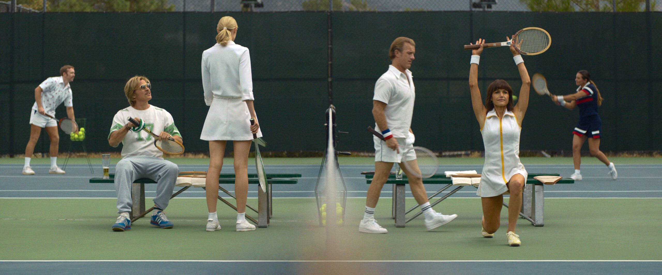 "People in white tennis outfits on a court; some are seated or standing by benches, one woman lunges with a racket raised, others play in the background on a bright outdoor court with a green fence."