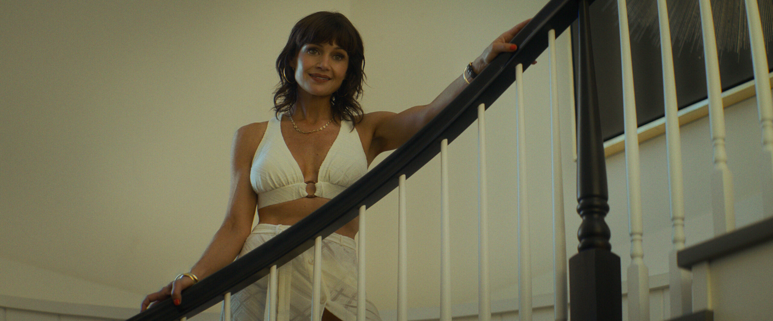 "A woman in a white outfit stands on a curved staircase indoors, smiling, with one hand on the railing. The setting has neutral walls and framed art, giving a modern and welcoming home atmosphere."