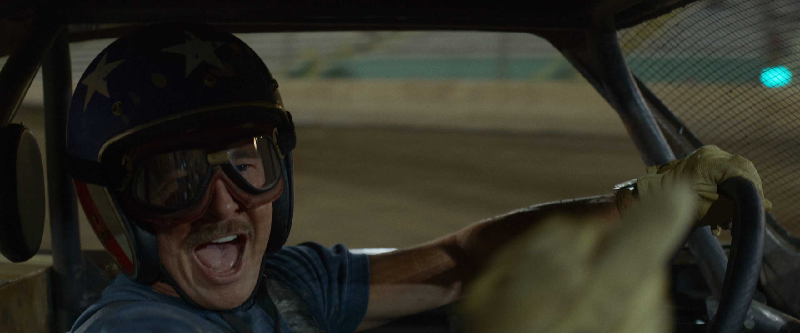 "Man wearing a helmet and goggles smiles excitedly while driving a race car on a dirt track, visible through the vehicle’s interior, suggesting a lively racing atmosphere."